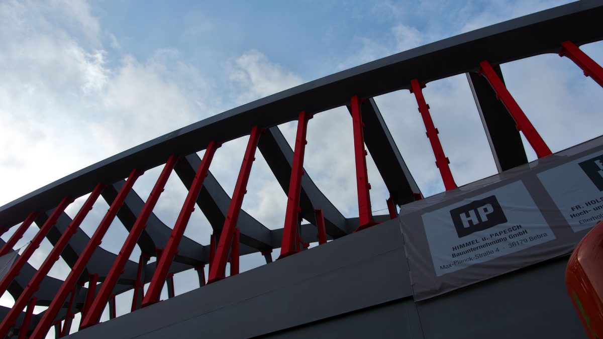 Red and black steel beams forming part of a modern architectural structure against a partly cloudy sky, with a construction company sign visible
